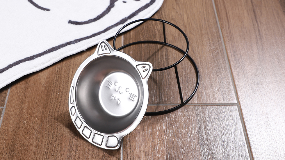 A small kitten sitting near two shallow stainless steel bowls on a washable mat in a quiet corner